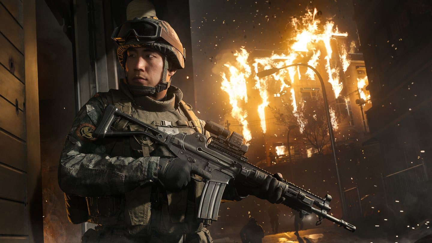 A young soldier in a helmet with goggles holds a rifle in a doorway, a building burning across the street behind him.